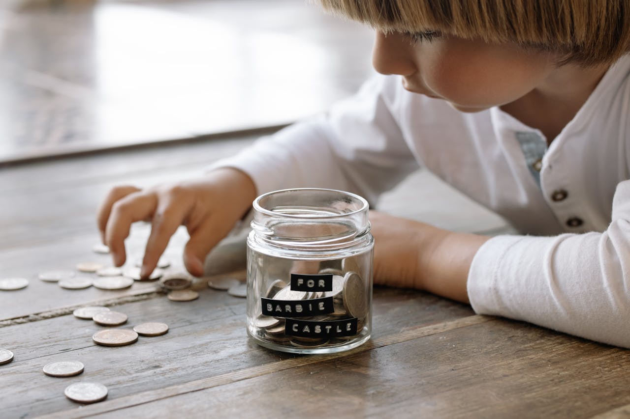 A young child collects coins in a jar labeled 'For Barbie Castle', symbolizing saving and dreams.